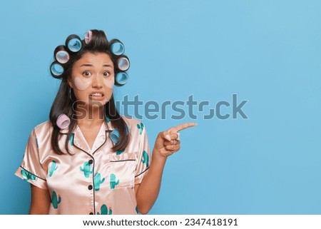 Pretty girl with curlers on her black hair points index finger aside, wearing silk pijamas, picture setup on blue background, good mood concept, copy space