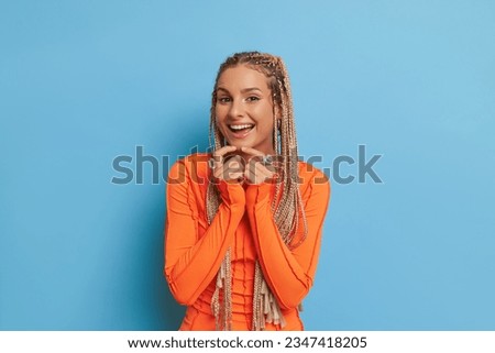 Closeup shot of cheerful girl with long dreadlocks wearing orange longsleeve top on blue background with hands pressing to her chest, good mood concept, copy space