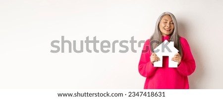 Real estate. Beautiful dreamy asian lady hugging paper house model with closed eyes, smiling as dreaming about buying apartment, standing over white background. Royalty-Free Stock Photo #2347418631