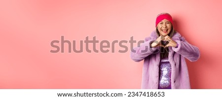 Love and fashion concept. Happy asian senior female showing heart sign and smiling at camera, I love you gesture, standing against pink background.