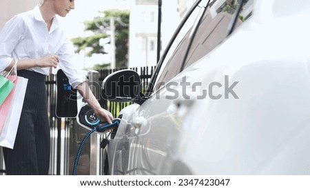 Businesswoman with shopping bag recharge her EV car display smart digital battery status hologram from charging station in car park or mall. Futuristic lifestyle and eco-friendly car. Peruse