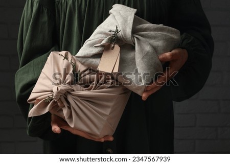 Furoshiki technique. Woman holding gifts wrapped in fabric with thuja branches near dark brick wall, closeup Royalty-Free Stock Photo #2347567939