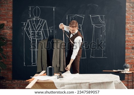 Stylish young man, fashion design in his atelier studio, making measurements of suit, creating classical style clothes. Concept of fashion, profession, creativity, occupation, hobby, business