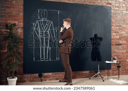 Young concentrated and thoughtful man, fashion designer standing by black board and attenticlelly looking at suit sketches. Concept of fashion, profession, creativity, occupation, hobby, business