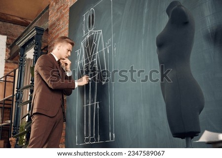 Thoughtful and concentrated young man, fashion design in classical clothes making suit sketches on blackboard. Concept of fashion, profession, creativity, occupation, hobby, business
