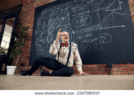 Young man, teacher, scientist sitting by blackboard with scientific calculations and formulas, finding out solution. Concept of education, science, profession and occupation, knowledge