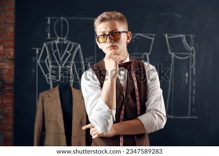 Handsome, stylish young man, fashion designer in classical clothes standing against blackboard with clothing sketches. Concept of fashion, profession, creativity, occupation, hobby, business