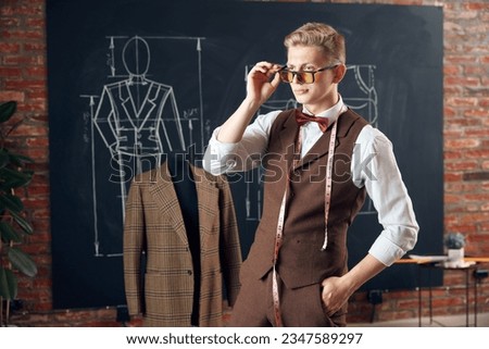 Handsome, stylish young man, fashion designer in classical clothes standing against blackboard with clothing sketches. Concept of fashion, profession, creativity, occupation, hobby, business