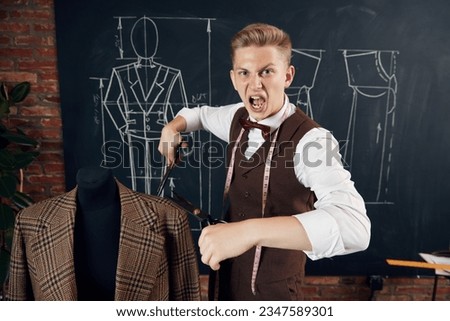 Emotional young man, fashion designer standing with scissors near mannequin with classical suit. Concept of fashion, profession, creativity, occupation, hobby, business