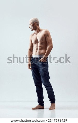 Full-length portrait of handsome young man with blonde hair, musuclar body posing shirtless in jeans against grey studio background. Concept of male natural beauty, body care, health, sport, ad Royalty-Free Stock Photo #2347598429