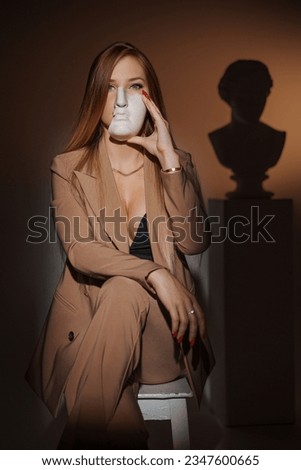 Beautiful woman in an elegant beige suit, partially hiding her face with a plaster ancient Greek mask against a warm illuminated background, with the silhouette of an ancient Greek bust visible