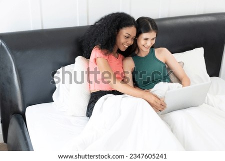 Happy lesbian couple using laptop on bed at home. Young multiethnic lesbian couple using laptop together in bedroom.