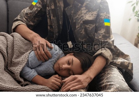 Soldier in Ukrainian military uniform kissing his daughter while she sleeping on sofa at home. Family reunion.