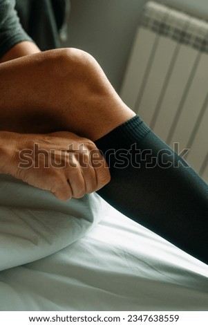 a man sitting on the bed puts on a black compression sock, with daylight