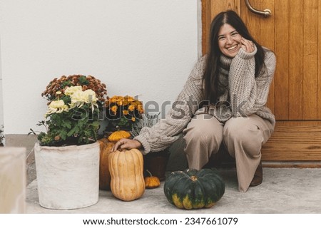 Happy woman in knitted sweater decorating house entrance with autumn pumpkins, pots with chrysanthemums and heather. Stylish autumnal decor of farmhouse front door. Fall arrangement