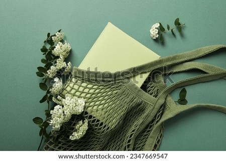 Book in string bag and flowers on green background, top view
