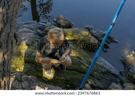 A boy sitting on rocks by the river equips the hook with bait. Sport fishing on the river in summer.