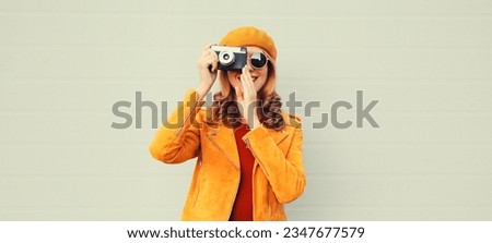 Fashionable autumn color style outfit, stylish young woman photographer with film camera wearing orange french beret hat, jacket, sunglasses on gray background