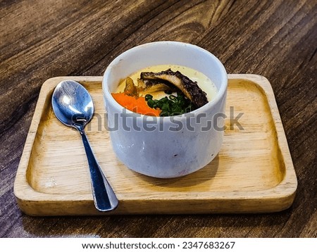 Japanese-style steamed egg topped with eel, shredded green onions and shrimp roe in a white cup on a wooden tray.