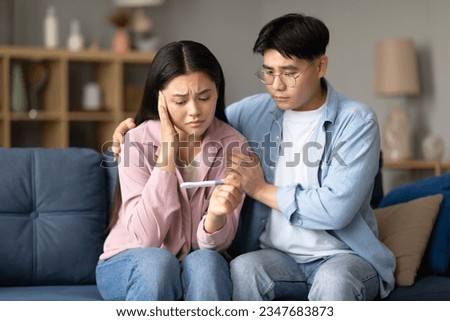 Childbirth Problem. Depressed Korean Couple Holding Negative Pregnancy Test, Sitting Together On Couch At Home. Young Spouses Struggling With Infertility Issue And Dreaming About Baby Royalty-Free Stock Photo #2347683873