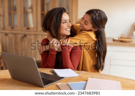 Happy Arab Little Daughter Embracing Mom While She Working Online On Laptop, Sitting At Desk At Home. Mother And Kid Girl Cuddling, Posing Near Computer. Internet Technology And Familly Life