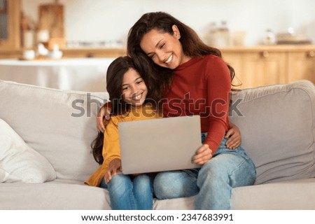 Happy Young Arabic Mother And Daughter Using Laptop Computer, Websurfing And Watching Cartoons Online Together Sitting On Sofa At Home. Mom And Kid Girl Browsing Internet Via Computer