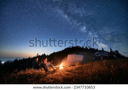 Night camping in the mountains under starry sky with Milky way. Travelling couple having a rest at campfire, tourist tent and off-road vehicle in the evening on mountain lawn and stargazing together. Royalty-Free Stock Photo #2347710853