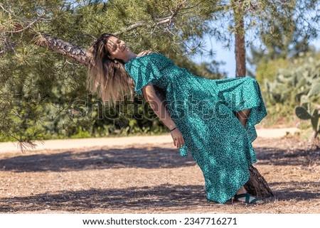 Cute blond girl with green dress leaning on a tree on a park.