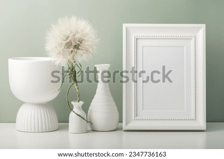 An empty photo frame and three white vases and a dandelion on the table. Minimalistic interior.