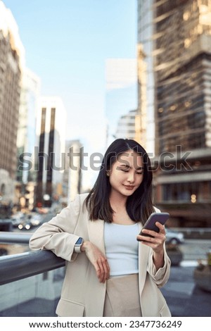 Young pretty Asian business woman office professional with cellphone in hand standing on big city street holding mobile device, using smartphone, looking at cell phone, texting messages, vertical. Royalty-Free Stock Photo #2347736249