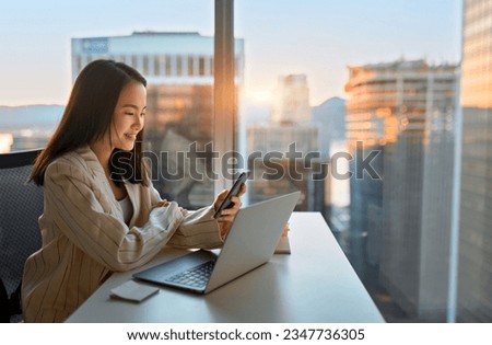 Young busy happy Asian business woman manager using mobile cell phone tech in office. Professional female executive holding smartphone, working, checking cellphone sitting at desk at big window.