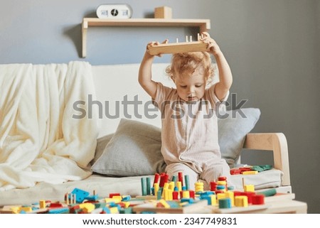 Nursery room filled with exploration. Building blocks of learning. Fun activities for young children. Funny wavy haired blonde infant baby raised arms with wooden educational toys at home interior. Royalty-Free Stock Photo #2347749831