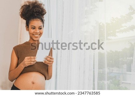 Happy portrait african american pregnant mother standing by home window looking at upcoming baby's ultrasound with fanciful anticipation and delighted to see healthy fetal evolution.