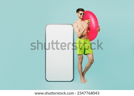Full body young man wear green shorts swimsuit relax near hotel pool hold ring big huge blank screen area mobile cell phone use smartphone isolated on plain blue background. Summer sea sun tan concept
