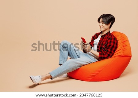 Full body young man of Asian ethnicity he wear red shirt casual clothes sit in bag chair hold in hand use mobile cell phone isolated on plain pastel light beige background studio. Lifestyle concept Royalty-Free Stock Photo #2347768125
