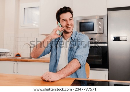 Young caucasian handsome guy having a friendly telephone call conversation sitting at home with positive expression. One young man smiling, laughing and talking by cell phone at kitchen room. High Royalty-Free Stock Photo #2347797571