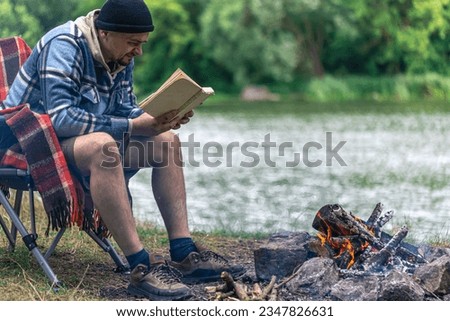 Close-up of bonfire with man reading a book in the background in nature.