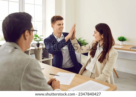 Happy young family couple sign purchase agreement at real estate agent's office. Joyful business partners make a deal. Smiling husband and wife buy house, sign contract and high five each other