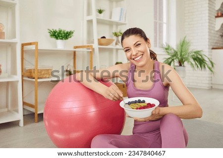 Portrait of a young sporty cheerful slim woman wearing sportswear eating healthy food after working out at home sitting on the floor on yoga mat. Healthy nutrition and lifestyle concept. Royalty-Free Stock Photo #2347840467