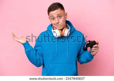 Young brazilian man playing with a video game controller isolated on pink background having doubts while raising hands