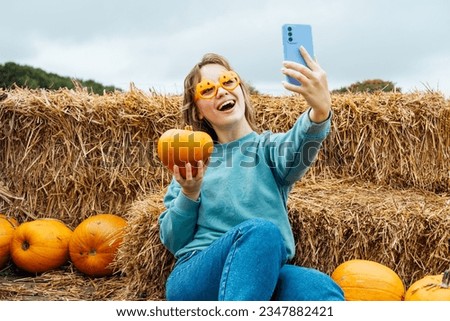 Smiling woman in fun glasses sitting on straw bales and holding pumpkin and making selfie on phone. Selecting Thanksgiving and Halloween holidays decor on agriculture farm. Autumn fall festive mood.