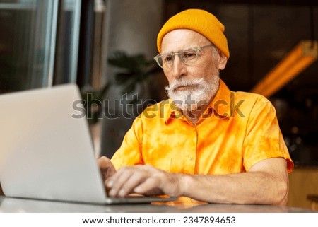 Handsome senior man, bearded programmer wearing yellow hat using laptop computer working online at workplace. Stylish freelancer, copywriter typing on keyboard sitting in modern cafe. Remote job