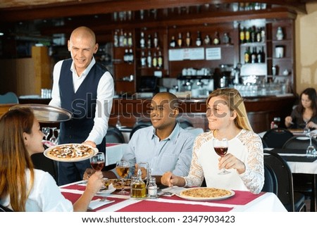 Portrait of polite smiling waiter serving pizza to friendly company in cozy pizzeria