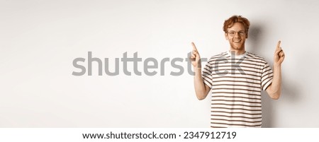 Funny young redhead guy in nerdy glasses pointing fingers sideways, showing two promo offers, standing happy over white background.