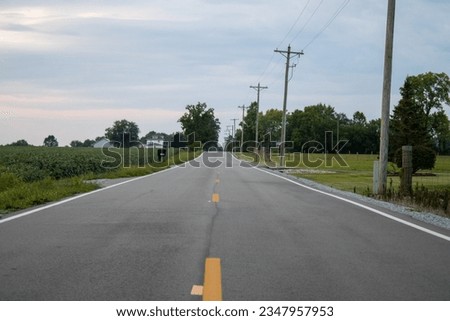 A rural road in Dayton Ohio with farm and bean fields and telephone poles with trees in the background and a house