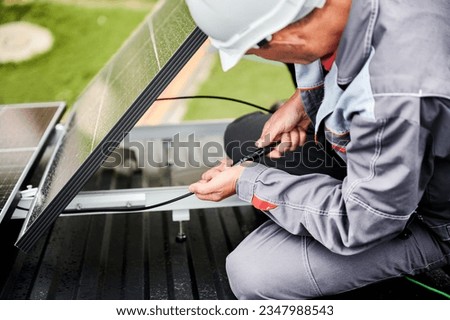 Professional specialist connecting cables of photovoltaic solar panel. Installation of pv solar cell on a top of house. Worker in white helmet securing cables of solar panel on a roof. Royalty-Free Stock Photo #2347988543
