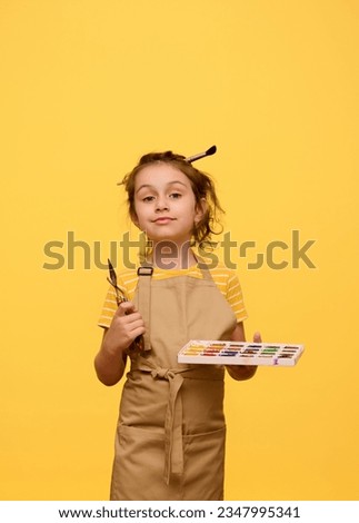 Caucasian adorable confident creative little child, primary school girl holding watercolor palette, paint brush and palette knife, looking at camera, isolated over yellow studio background. Fine art
