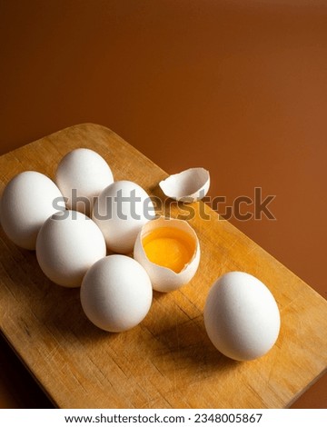 White chicken eggs on yellow wooden board on dark orange background. One open egg with visible yolk next to whole eggs in the kitchen. Ingredients for cooking on bright background. Vertical photo