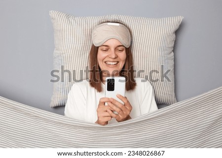 Happy charming woman in white T-shirt and sleeping eye mask lie in bed on pillow under blanket isolated on gray background using cell phone reading internet news smiling happily
