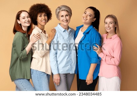 Group of beautiful smiling multiracial women wearing stylish colorful shirts looking at camera isolated on beige background. Portrait happy different ages fashion models posing for pictures in studio 